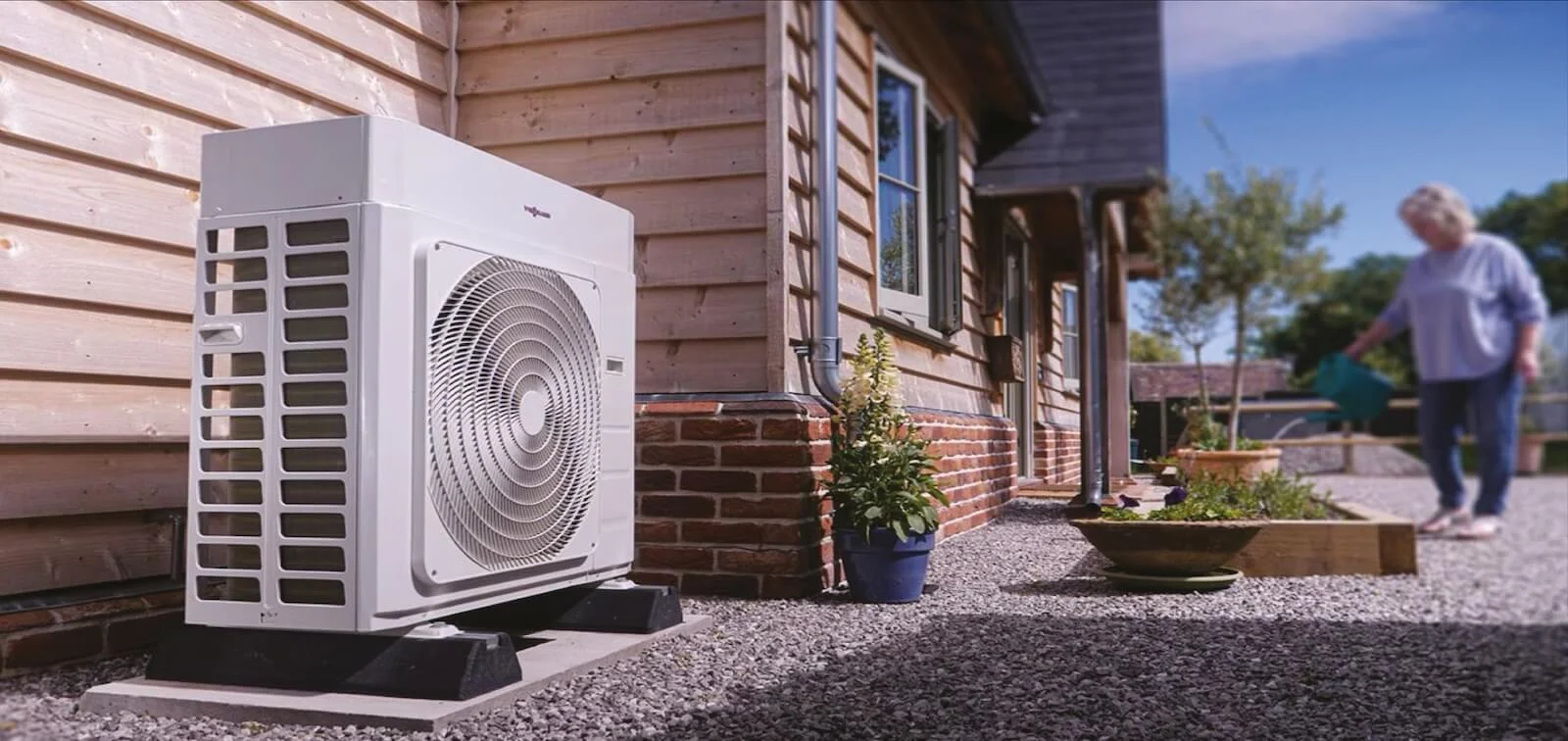 An outdoor heat pump unit is installed next to a wooden house on a gravel path. A potted plant is nearby, and a person in a sweater walks towards raised garden beds in the background, under a clear blue sky.