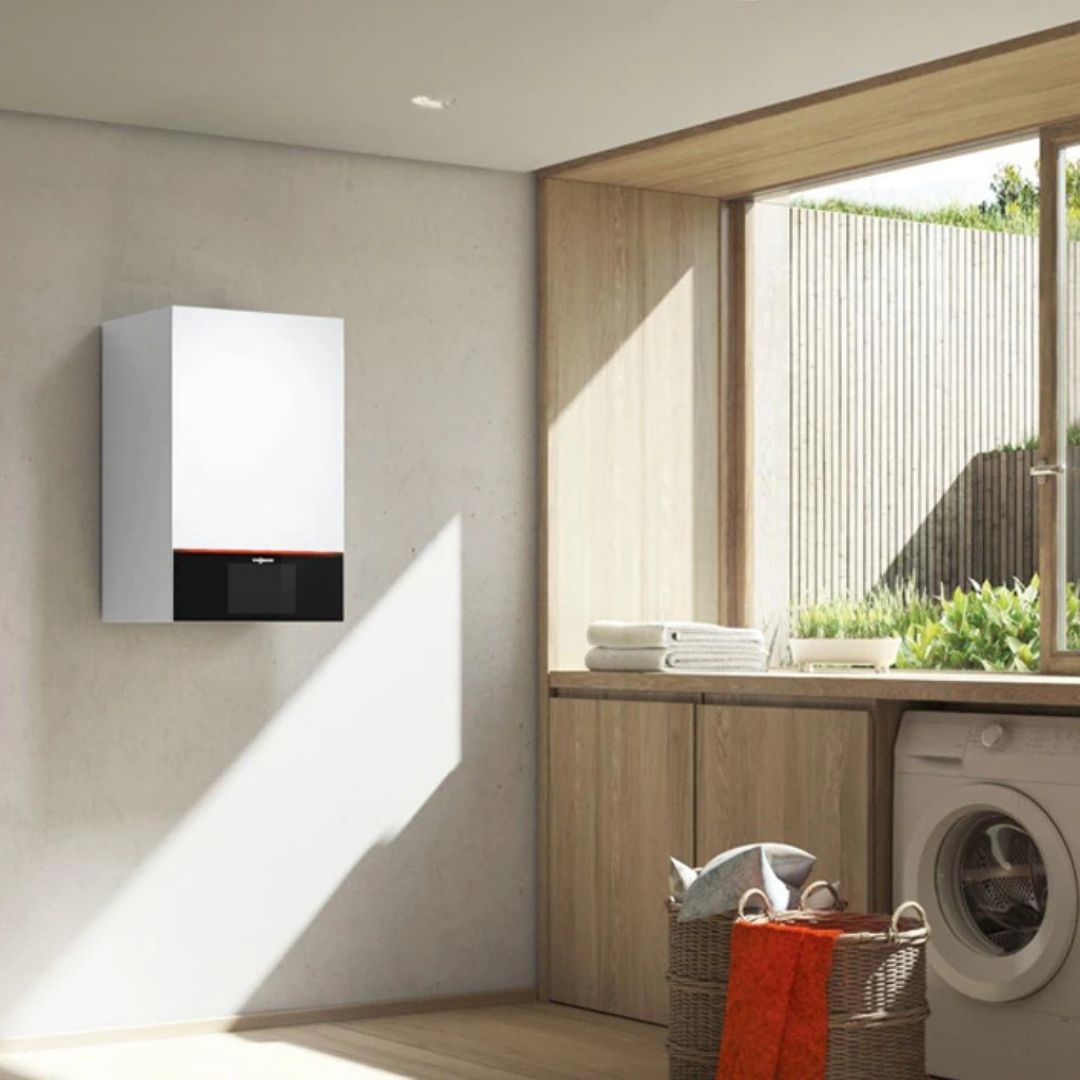 Modern laundry room with a wall-mounted boiler, wooden countertops, and a washing machine. Sunlight streams through a large window illuminating folded towels and a red basket on the counter. Outdoor greenery is visible through the window.