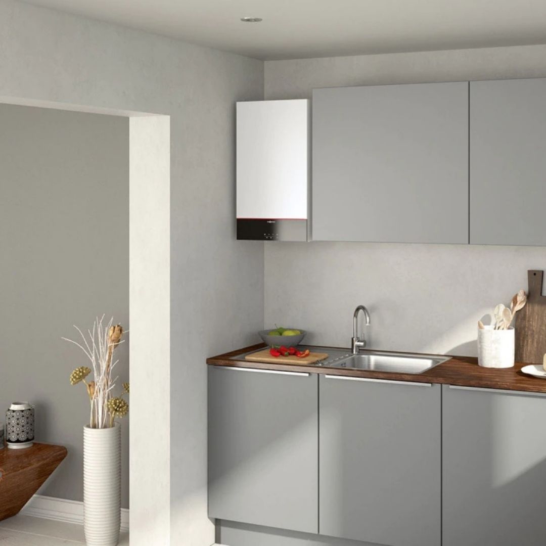 A modern kitchen features light gray cabinets with a wooden countertop. A white wall-mounted appliance is above the counter, and a sink with a faucet is visible. A vase with flowers stands in the corner, and a bowl of fruit is on the counter.