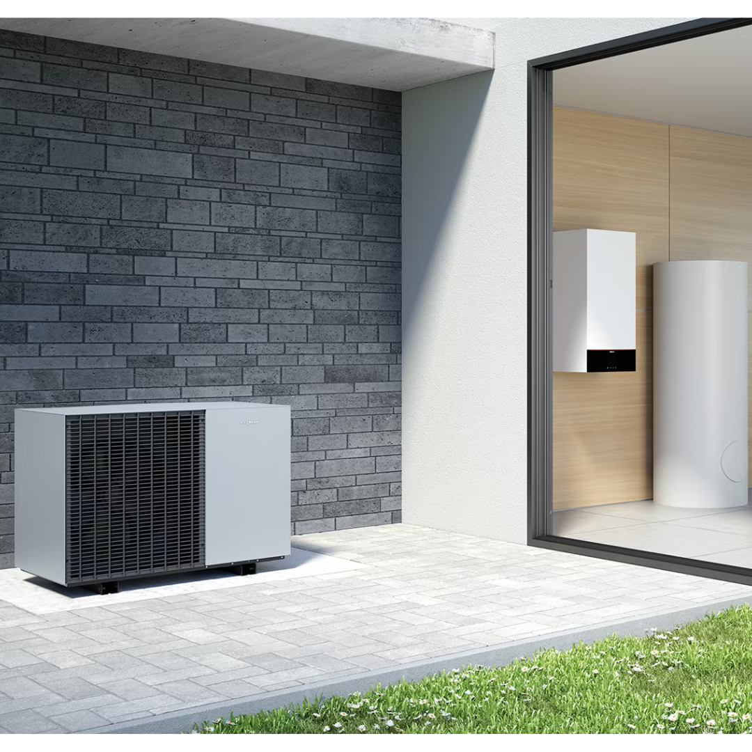 A modern heat pump system is installed outside a contemporary building. The unit sits on a paved area next to a grass lawn, with a view into a room where additional equipment is mounted on a wooden wall.