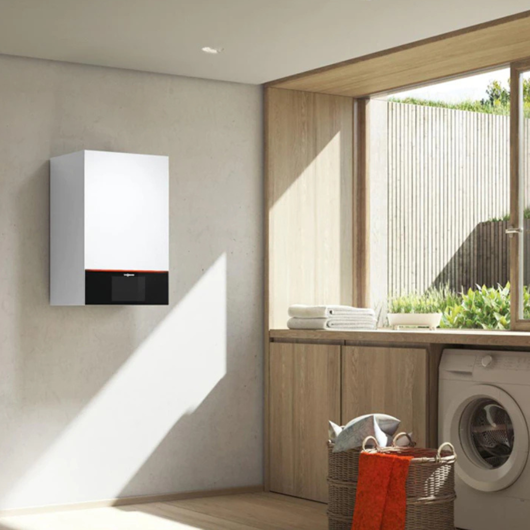 A modern laundry room features a wall-mounted boiler from a recent boiler installation, washing machine, and neatly folded towels on a wooden counter. Natural light floods through a large window, highlighting a red cloth in a basket and underscoring the efficiency of the heating system.